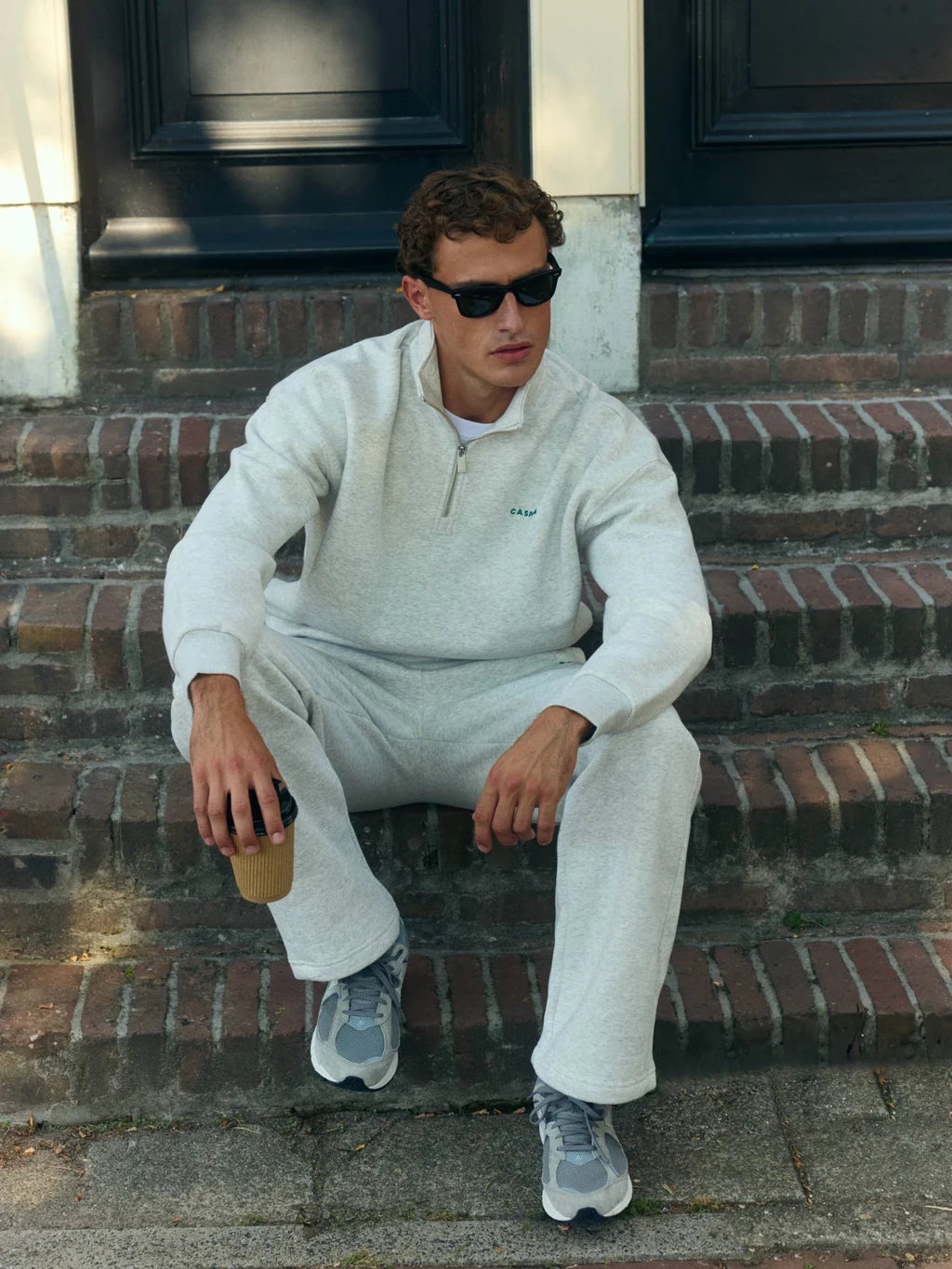 Man wearing a grey melange CASPARA quarter-zip sweatshirt sitting on outdoor brick steps.